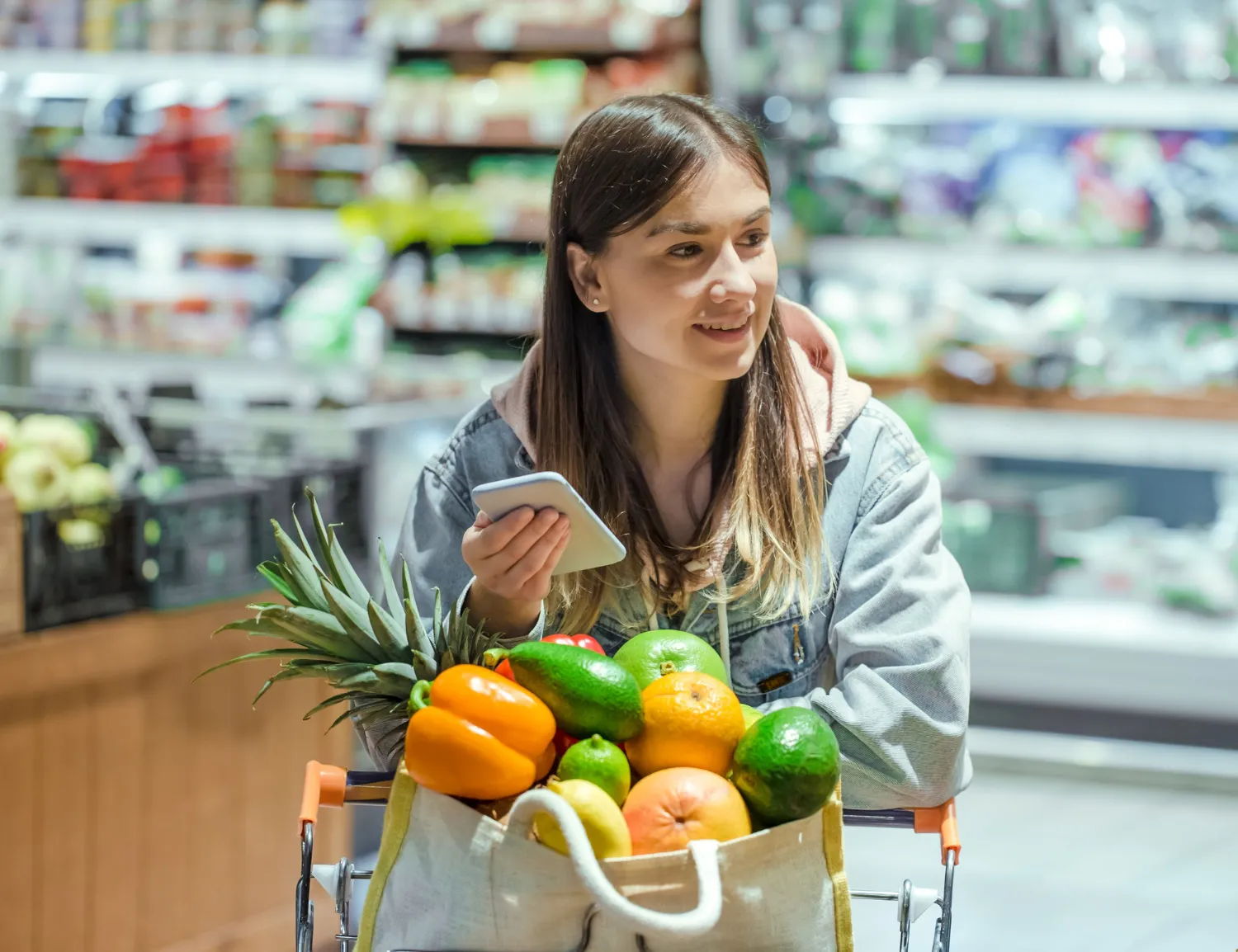Klientka z telefonem i wózkiem pełnym warzyw w alejce supermarketu